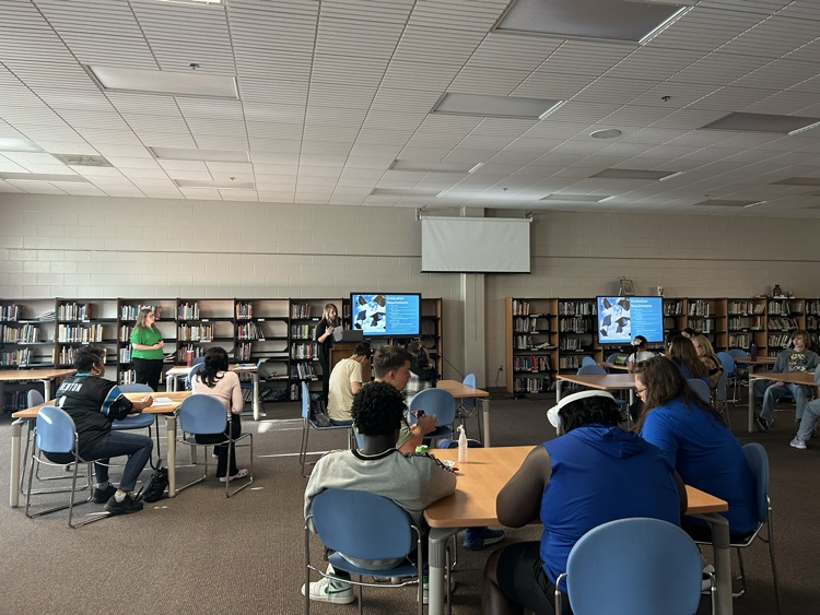 several people sitting at tables in a library looking at two screens while a woman presents information