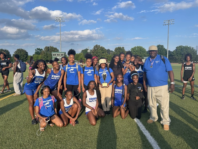 A high school track team poses on a field with their coach. Athletes wear blue uniforms, and one student holds a trophy. Stadium lights and trees are visible in the background.