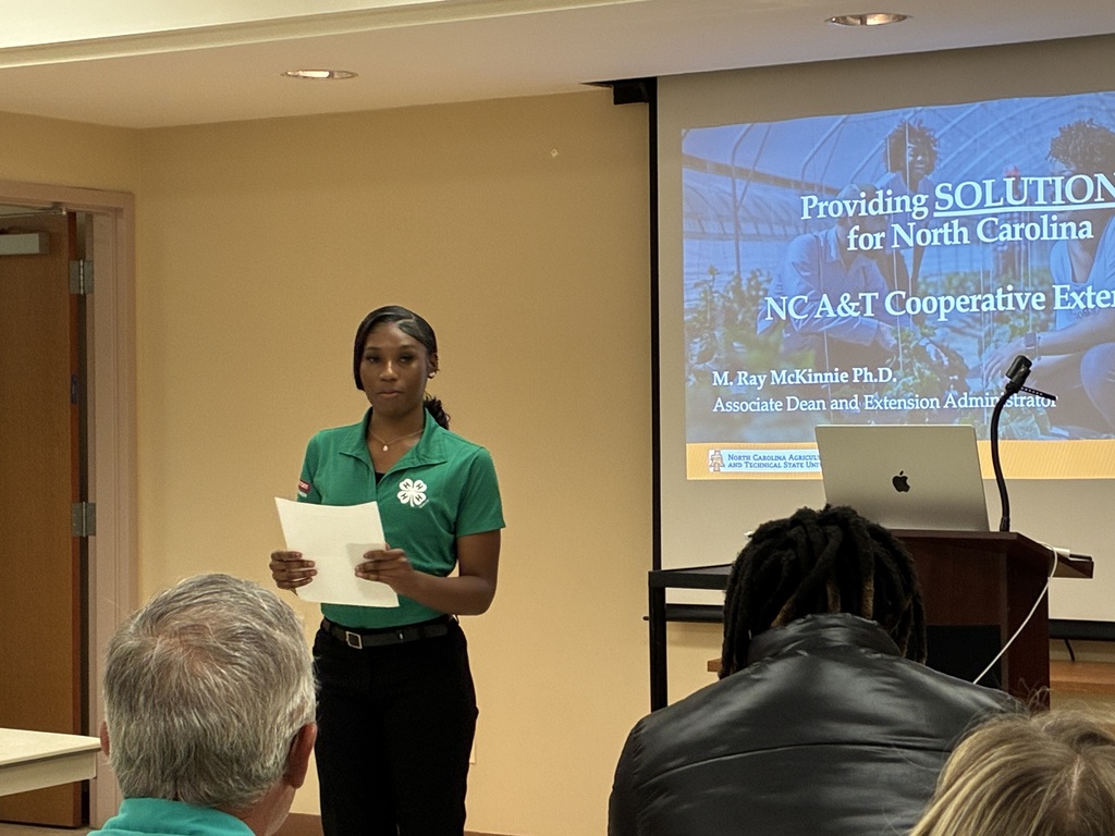 A young woman wearing a green 4-H polo shirt stands at the front of a conference room, holding papers and speaking to an audience. Behind her, a projector screen displays a presentation about NC A&T Cooperative Extension and solutions for North Carolina. Several attendees are seated in the foreground, facing the speaker, while a podium with a laptop and microphone stands beside the screen.