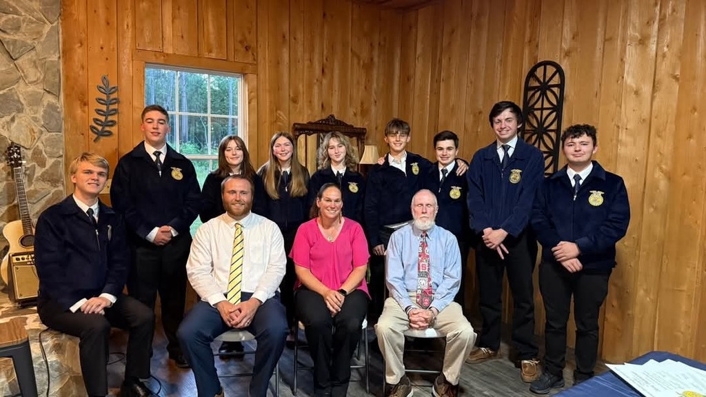 A group of nine West Bladen High School students and two adults in formal FFA attire and business casual wear pose together in a rustic wood-paneled room with a stone fireplace.