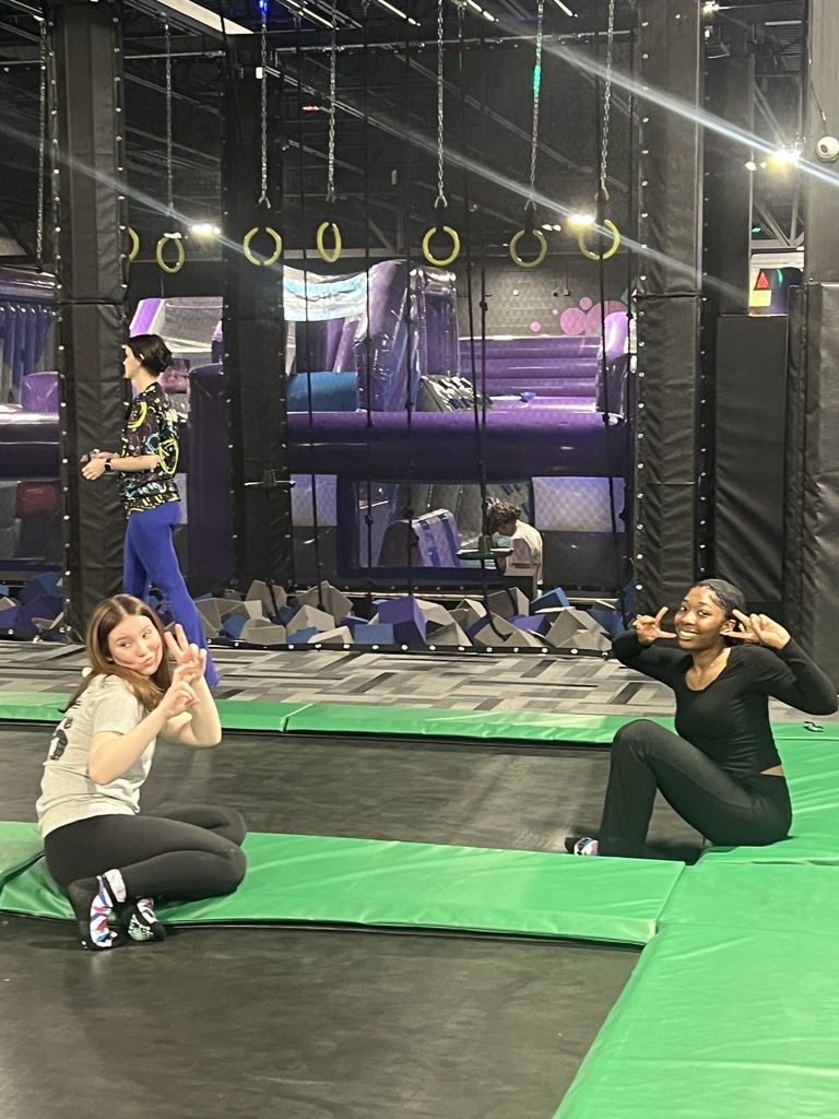 Two young women smiling and posing with peace signs in the foreground of an indoor trampoline park. In the background, other children play on purple and black padded equipment with hanging rings and foam pits.