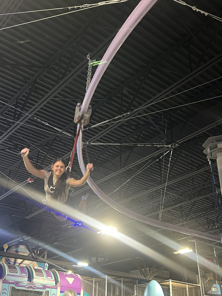 A student smiles and raises her arm while being suspended in a safety harness from the ceiling of an indoor adventure park. Metal rafters and cabling are visible above her.