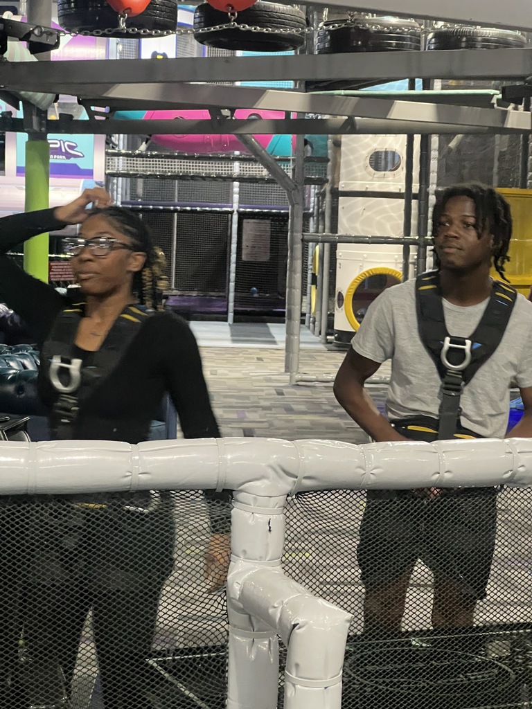 Two students at an indoor adventure park wearing safety harnesses and preparing for an activity. One student adjusts her hair while the other looks toward the side, with a climbing structure and padded obstacles in the background.
