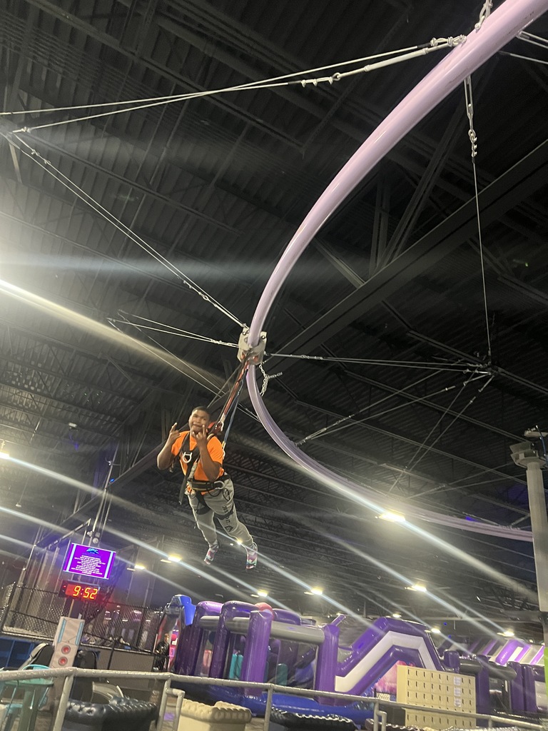A student wearing a safety harness smiles while being hoisted by ropes toward the ceiling of an indoor adventure park. Metal rafters and safety cables are visible in the background.