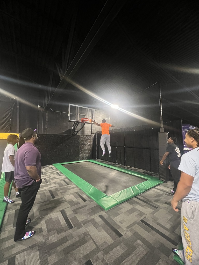 A student in an orange shirt jumps toward a basketball hoop on a trampoline at an indoor sports park. Several other students watch from the padded green floor below under bright overhead lights