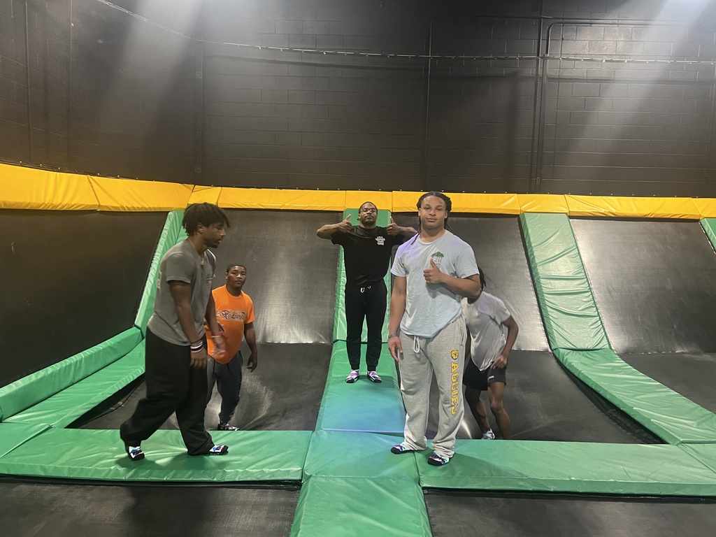Four male students posing on green padded mats at an indoor trampoline park. One student in the foreground gives a thumbs-up, while others stand behind him near angled trampolines with yellow and black safety padding