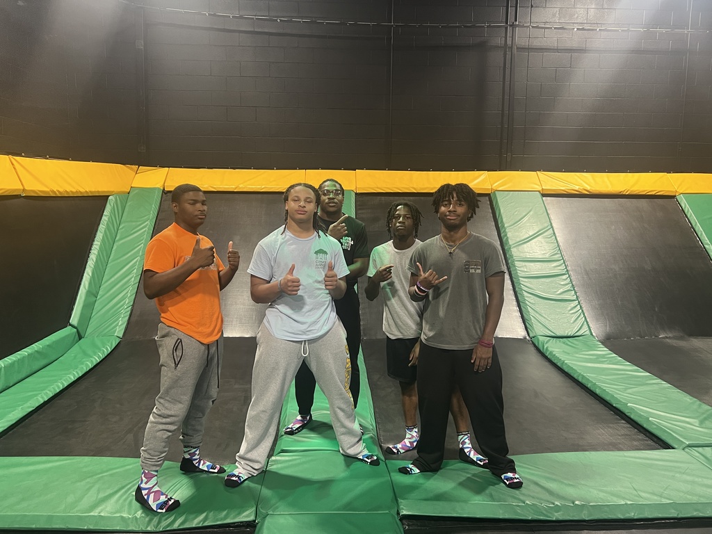 Four male students smiling and posing together on a green padded floor at an indoor trampoline park. They are standing in front of angled trampolines with yellow and black safety padding