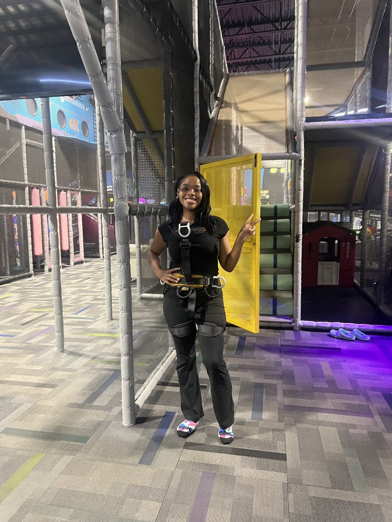 A student wearing a safety harness stands and smiles inside an indoor adventure park. Behind her is a yellow gated entrance to a climbing structure and various padded play areas