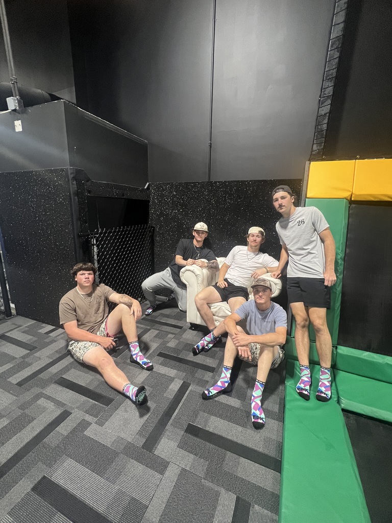 Five male students posing together on a white couch and the floor inside an indoor adventure park. They are in front of a black wall and green safety padding, with one student standing and the others sitting
