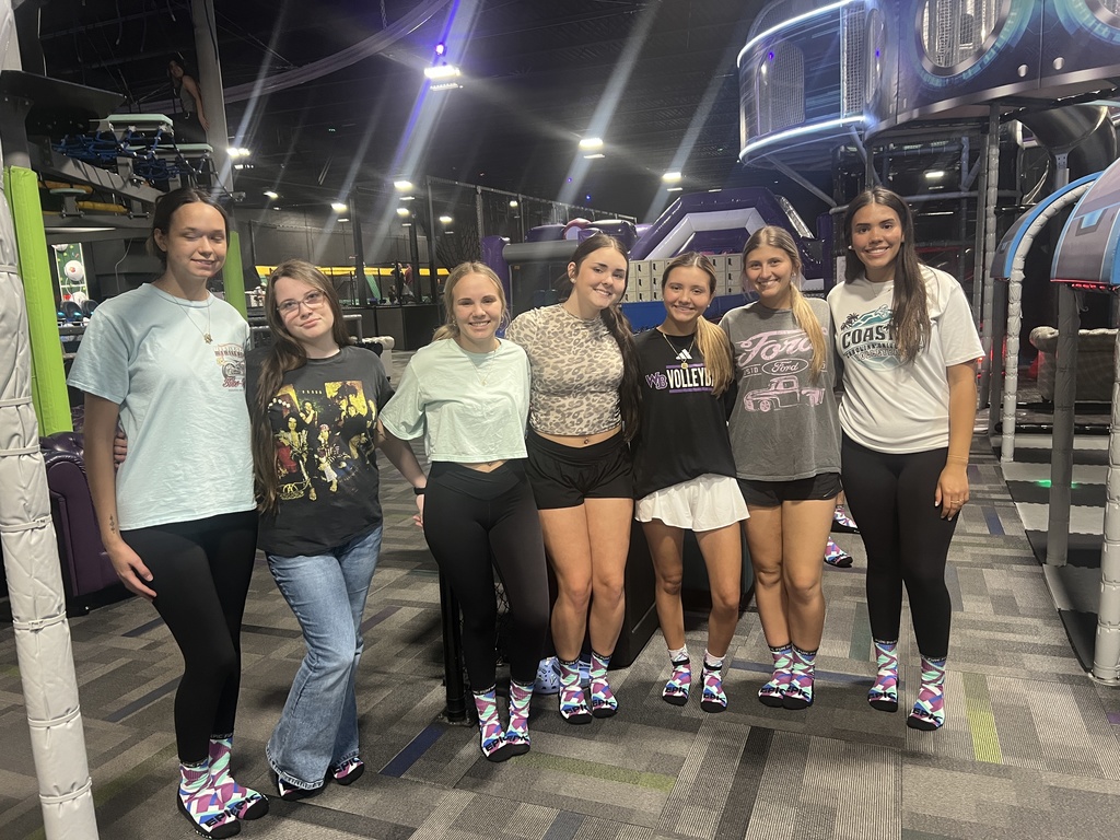 Seven female students smiling and posing together for a group photo at an indoor adventure park. They are standing on a carpeted floor in front of play structures and safety padding
