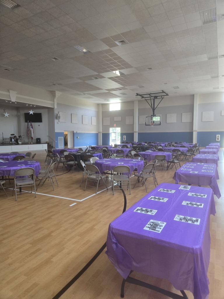An indoor event space at Galeed Church with several long tables covered in purple tablecloths, prepared for a dinner showcase.