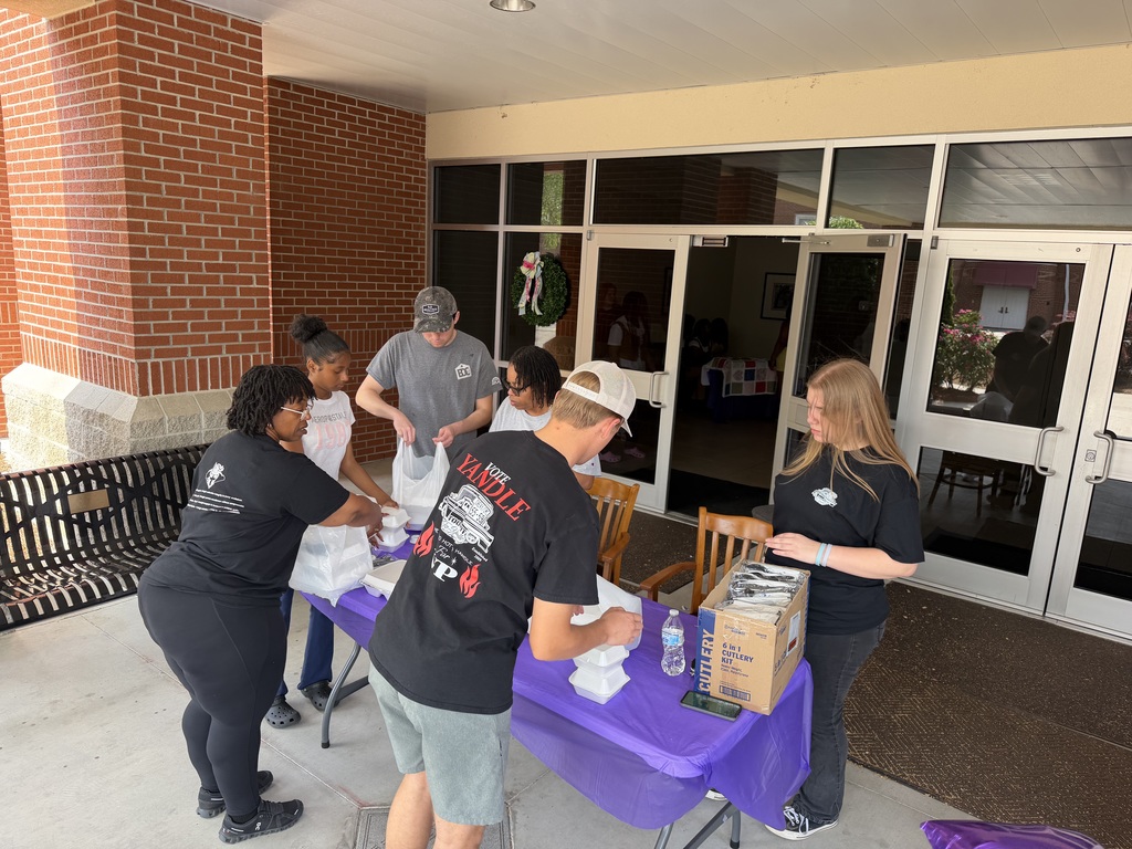A group of students and adults gathered under a building overhang, engaged in a conversation preparing to serve drive up guests