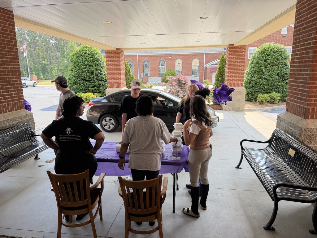 A group of students and adults gathered under a building overhang, engaged in a conversation with a parked black car in the background
