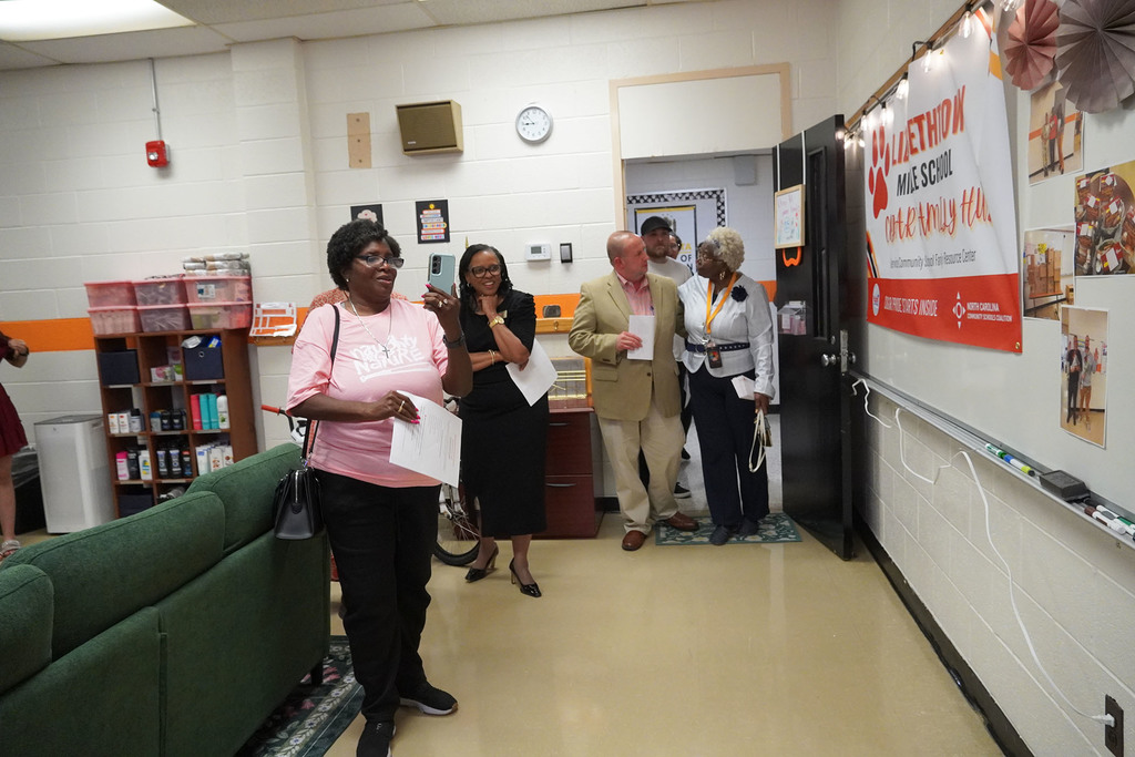 A group of adults walk into a room. You can see a green sofa, a book shelf with supplies, and photos on a white board. 