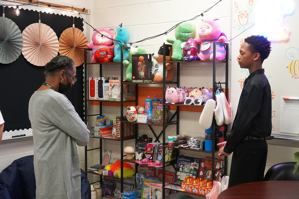 A student is talking with an adult. They are standing near shelves that have a variety of toys displayed.