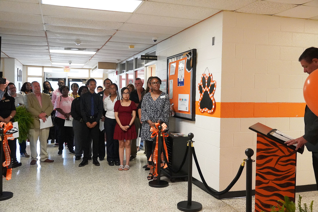 A group of people stand together listening to a gentleman speak from an orange and black podium. 