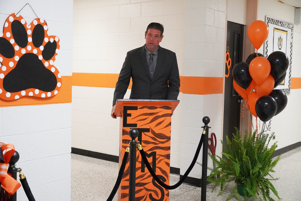 A school principal stands at an orange podium. You can see orange and black balloons beside him with a fern.
