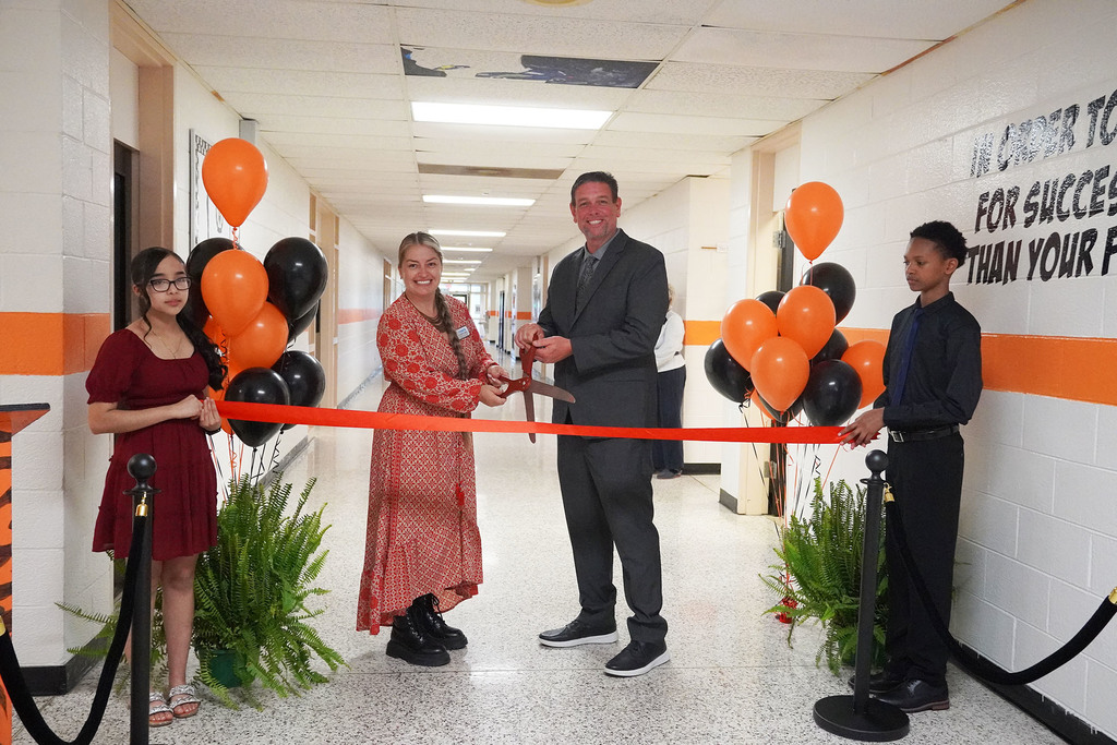 Two middle school students stand on opposite sides of the hallway holding a ribbon, while two adults stand with large scissors ready to cut.