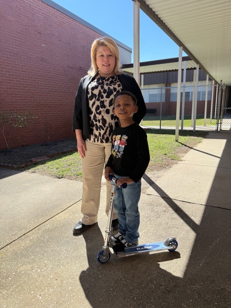 A school staff member stands outside with a smiling young student riding a scooter on a sunny day along a sidewalk beside a brick school building.