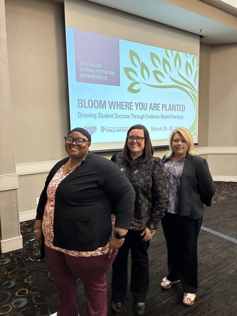 Three educators stand together smiling in front of a presentation screen at a conference that reads “Bloom Where You Are Planted: Growing Student Success Through Evidence-Based Practices.”