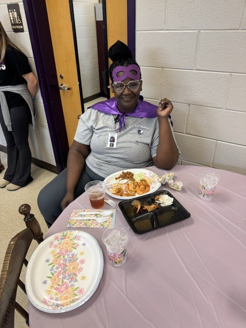 A woman is seated at a small table in what appears to be a school hallway or classroom area. She is smiling gently and posing with one hand raised near her face in a relaxed, playful way. She is wearing a gray shirt with a name badge clipped to it and a purple superhero-style mask, along with clear-framed glasses. Her hair is styled up in a high bun, and a purple cape is tied around her shoulders, matching the mask.