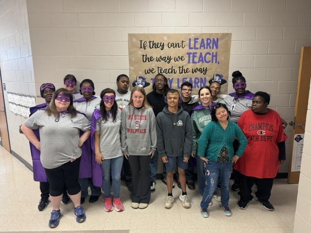 A group of about 14 students and adults are standing together in a classroom, posing for a group photo. They are arranged in two rows—some standing in front and others behind. Many of the individuals are wearing purple superhero-style masks and a few have matching purple capes, giving the group a fun, team-like appearance.