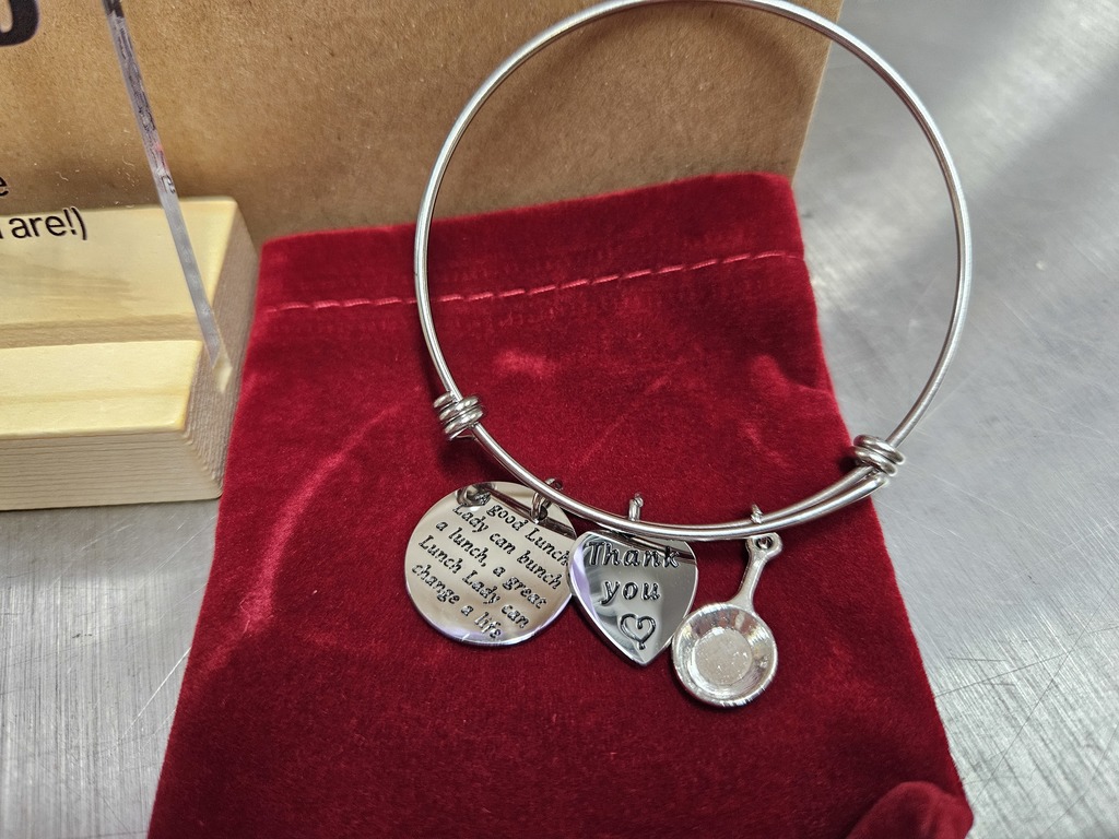 A silver wire bangle bracelet is displayed on top of a soft, red velvet pouch. The bracelet has several small charms attached near the front.  One charm is a round, flat disc engraved with a message about lunch, suggesting appreciation for someone who provides or supports meals. Next to it is a heart-shaped charm that clearly reads “Thank you” with a small heart symbol beneath the words. Another charm is shaped like a small frying pan, adding to the food or kitchen theme.  The bracelet and charms have a polished, reflective finish. The red velvet pouch beneath them provides a rich, soft background, making the silver jewelry stand out. To the left, part of a small wooden display stand is visible, and the surface underneath everything appears to be a smooth, metallic countertop.