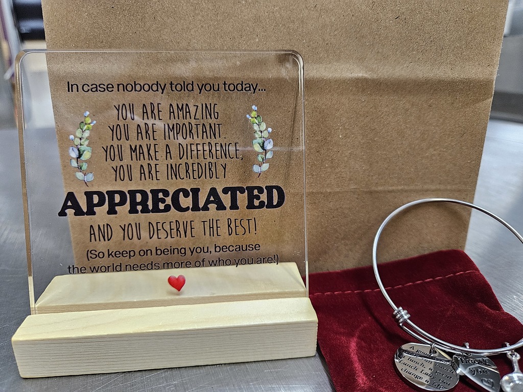 A small appreciation display is arranged on a metallic countertop. On the left, a clear acrylic sign stands upright in a light wooden base. The sign contains an encouraging message in mixed fonts that reads: “In case nobody told you today… You are amazing You are important You make a difference, You are incredibly APPRECIATED and you deserve the best! (So keep on being you, because the world needs more of who you are!)” The word “APPRECIATED” is printed in large, bold letters, making it the focal point. Simple leafy designs decorate both sides of the text, and a small red heart sits centered at the bottom of the wooden base. To the right of the sign, a silver wire bangle bracelet rests on a soft red velvet pouch. The bracelet has several charms, including a round engraved charm with a message about lunch, a heart-shaped charm that says “Thank you,” and a small frying pan charm, suggesting a food or kitchen appreciation theme. The overall scene feels warm and thoughtful, combining a heartfelt message with a small gift, likely intended to show gratitude to someone who helps prepare or provide meals.