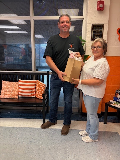 Man receiving a gift bag from a woman in a school hallway with orange and black decor.