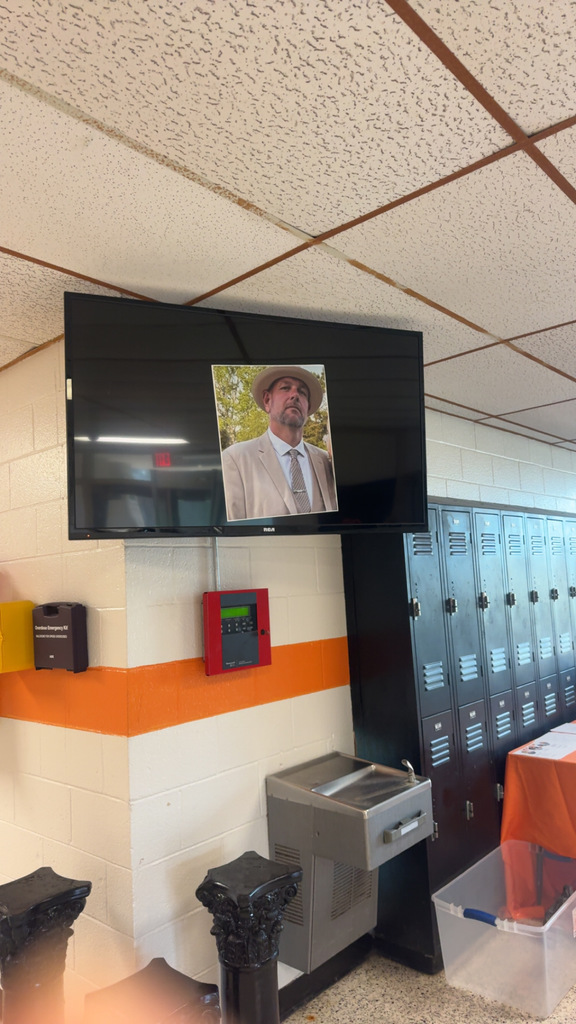School hallway with lockers and a TV screen displaying a portrait of a man in a tan suit and hat.