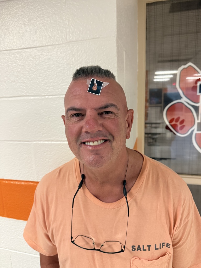 Smiling man wearing an orange shirt with glasses around his neck and a small photo sticker on his forehead.