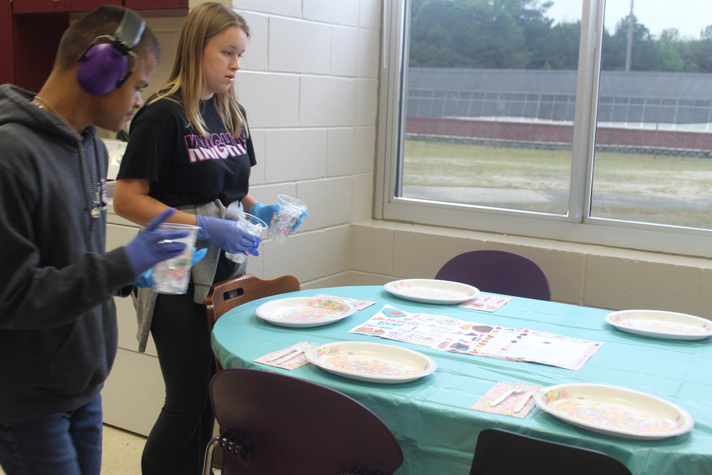 Two students from West Bladen High School, wearing blue gloves, carefully set a dining table with blue cloths and white plates in a classroom. In the background, a large window shows a view of the school grounds