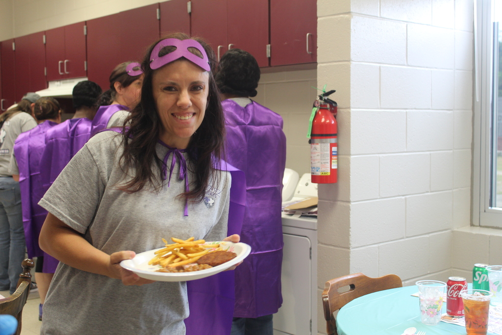 A school cafeteria worker smiling and holding a plate of food, including a burger and fries. She is wearing a gray shirt, a purple superhero cape, and a pink superhero mask. In the background, other staff members in superhero costumes are visible in a school cafeteria setting