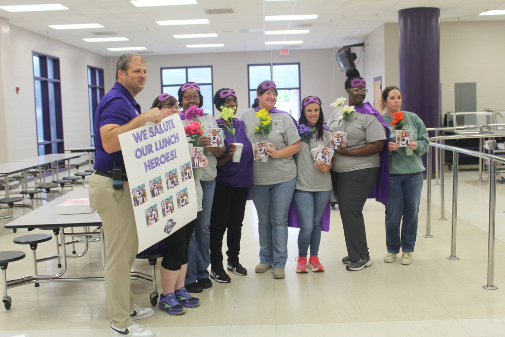 A group of six school staff members and a male administrator posing in a school cafeteria for School Lunch Hero Day. The staff members are wearing purple superhero capes and masks. The administrator on the left holds a large handmade sign that reads, "We Salute Our Lunch Heroes!" The group is smiling and standing in front of cafeteria tables and large windows
