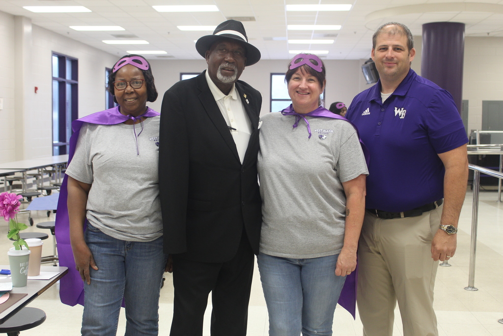 A group of four adults posing for a photo in a school hallway. Two women in the center wear matching gray t-shirts, purple superhero capes, and purple eye masks. They are flanked by a man on the left in a black suit and fedora, and a man on the right in a blue polo shirt. All four are smiling and standing close together.