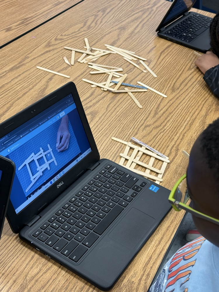 Student uses a Chromebook to follow a bridge-building design while assembling craft sticks on a classroom table.