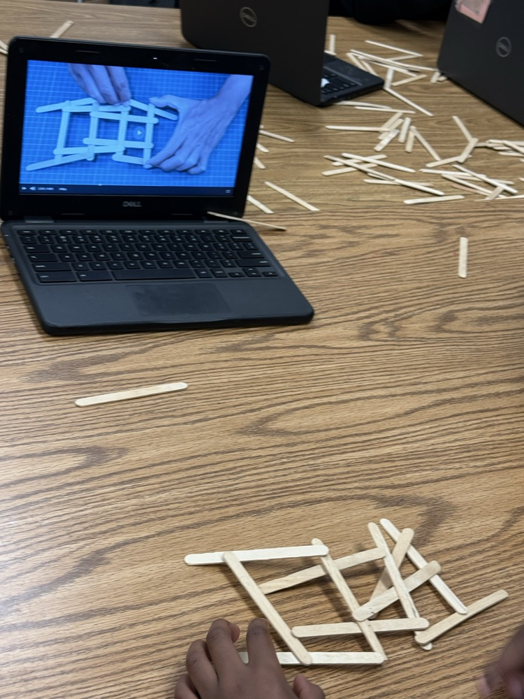 Student uses a Chromebook to follow a bridge-building design while assembling craft sticks on a classroom table.