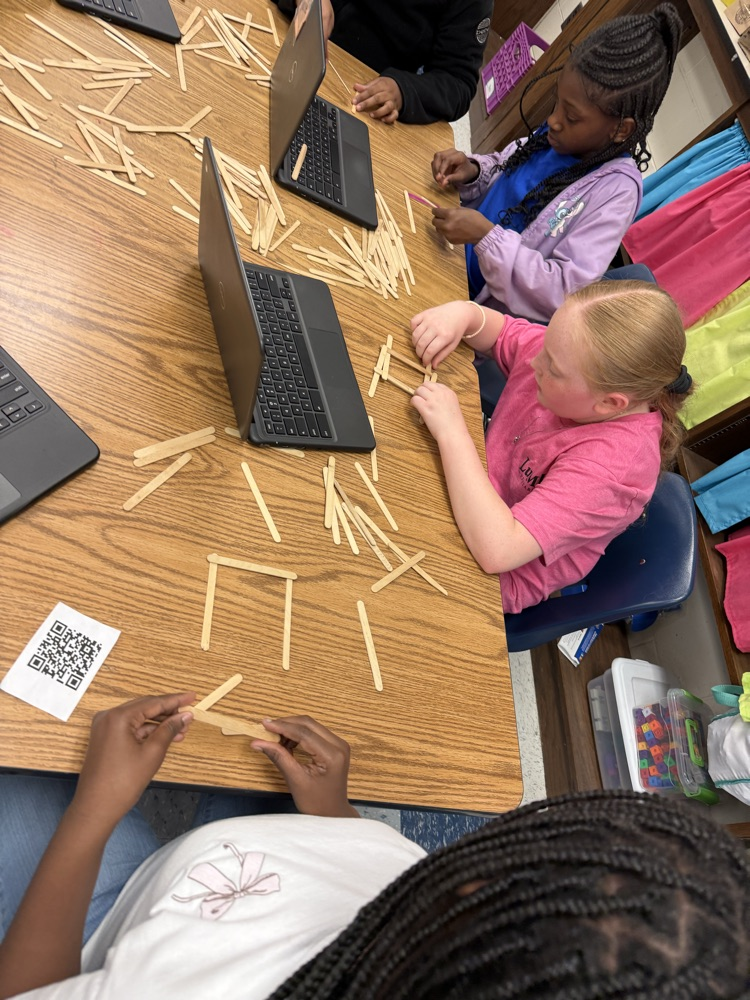Overhead view of students working with scattered craft sticks and Chromebooks while constructing bridge designs