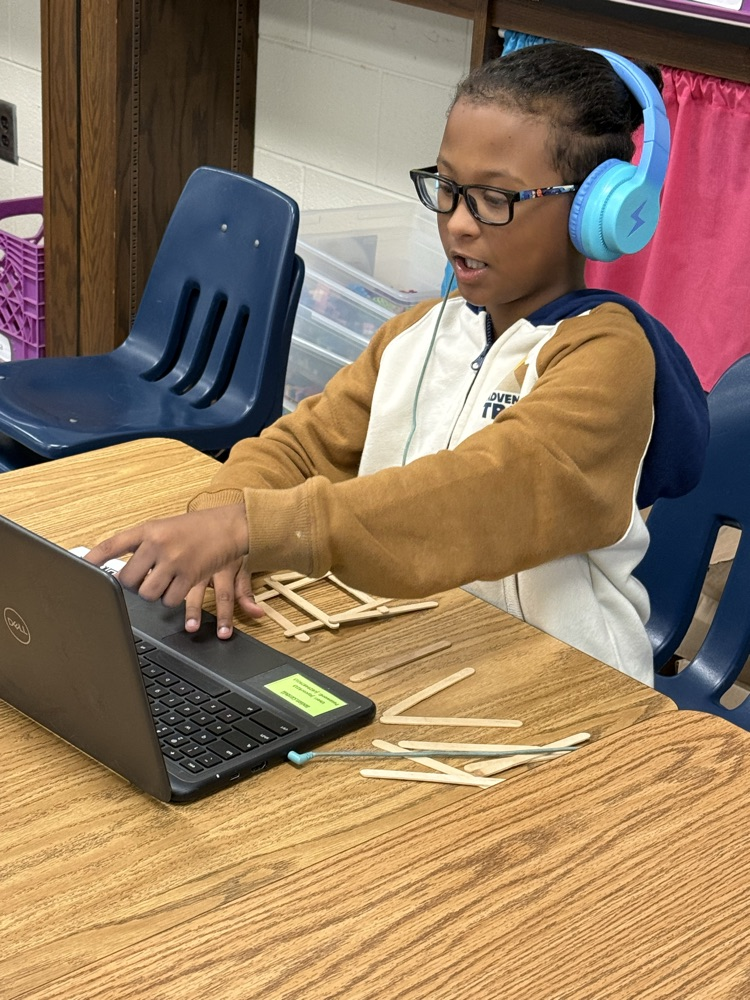 Student wearing headphones and glasses follows instructions on a Chromebook while building a bridge with craft sticks.