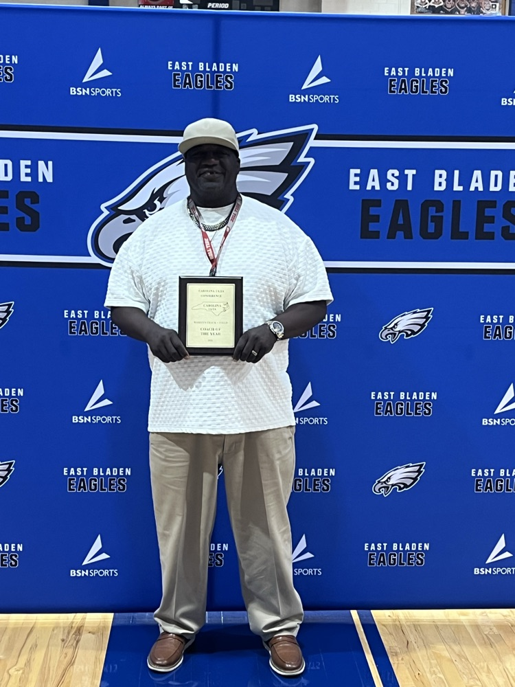 coach Wilson Bolden standing in front of East Bladen Eagles backdrop with eagle on it holding a plaque that shows he is the coach of the year for women’s track and field