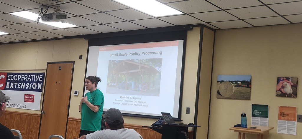 A student standing at a podium in a presentation room at the Bladen County Cooperative Extension office. A large projector screen behind her displays a presentation titled "Small-Scale Poultry Processing," featuring a photo of a chicken coop