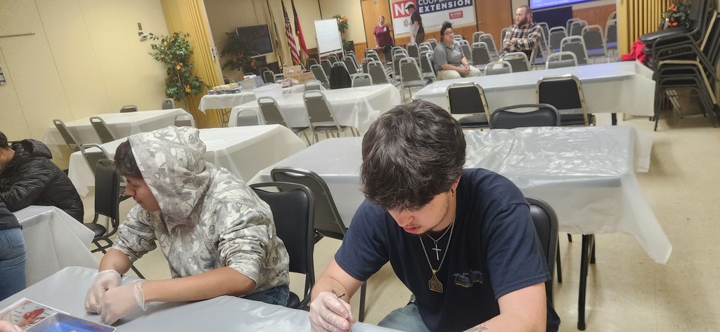 Two West Bladen High School students in a professional kitchen or processing environment work at a stainless steel table. They are wearing protective gloves and focusing on their task, with a large, organized meeting room visible in the background.