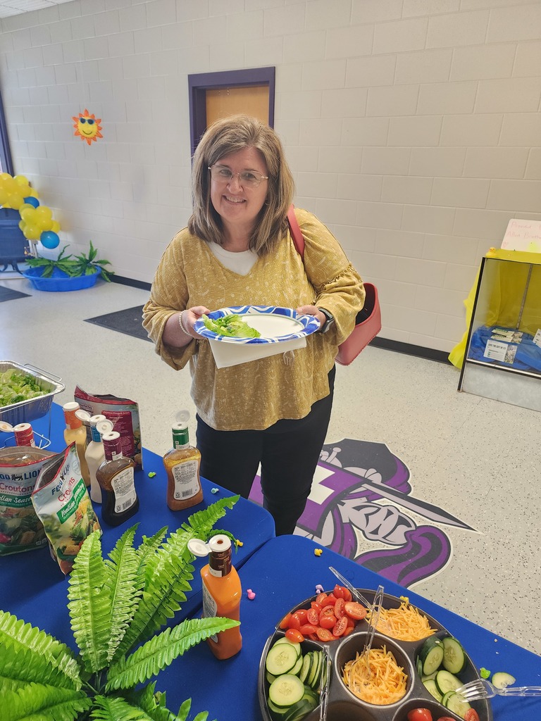 A woman smiling and holding a plate of salad at a catered school event. She stands behind a blue table featuring various salad dressings, toppings, and a purple West Bladen High School logo mat.