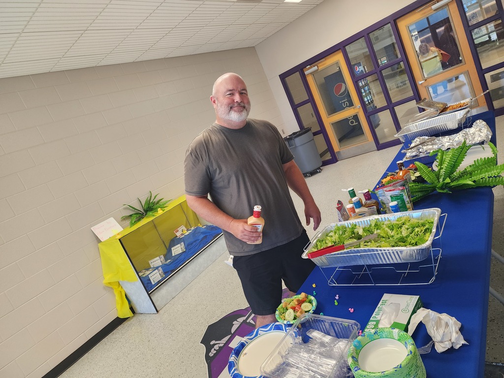 A smiling man holding a bottle of dressing stands behind a catered salad bar. The spread includes large bowls of lettuce, various toppings, and plates, set up inside a school building.