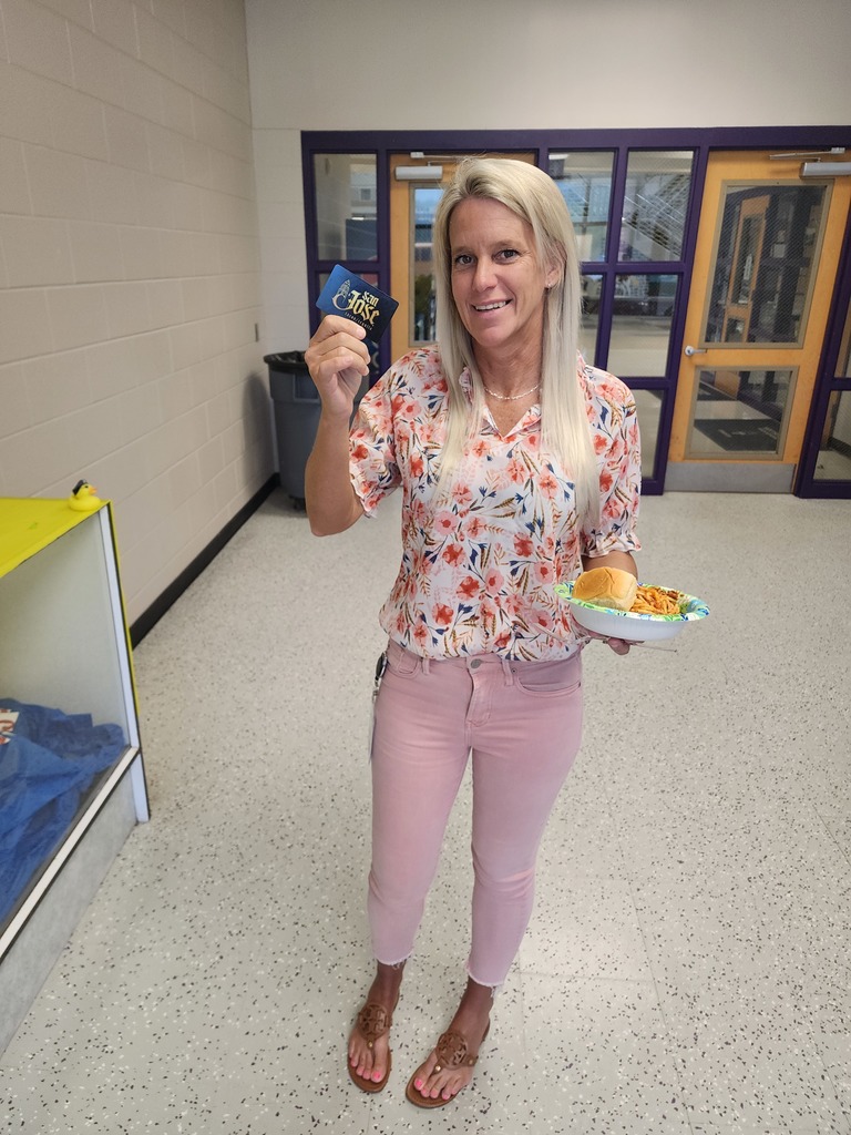 A woman in a floral blouse and pink pants smiles while holding a plate of food and a blue gift card. She is standing in a school hallway near a doorway.