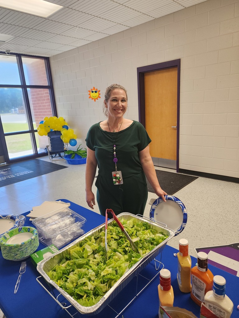 A female staff member with long brown hair and a green shirt smiling in a school hallway.