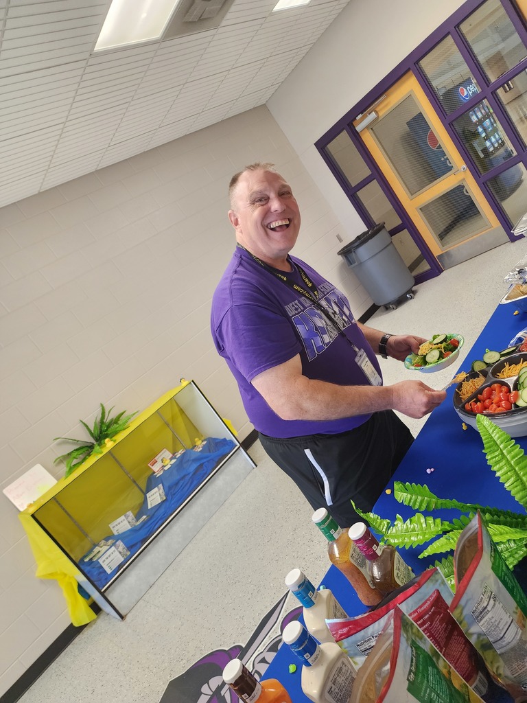 A smiling man in a purple t-shirt stands at a catered salad bar, holding a plate of food. He is positioned in a school hallway near a blue-draped table with various fresh vegetable toppings.