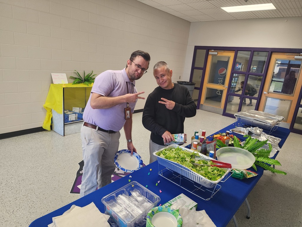 A smiling man holding a bottle of dressing stands behind a catered salad bar. The spread includes large bowls of lettuce, various toppings, and plates, set up inside a school building.