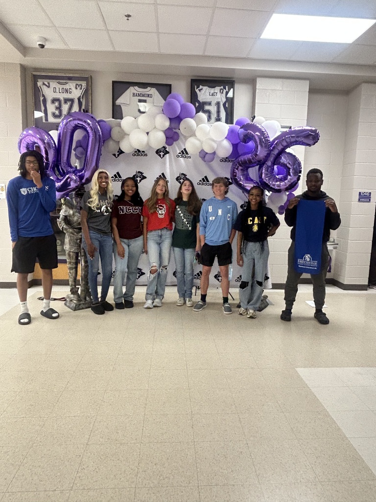 A group of students stand in a line in front of the Decision Day backdrop with purple and white balloons and “2026” balloon numbers. Some wear college shirts, showing their future plans.