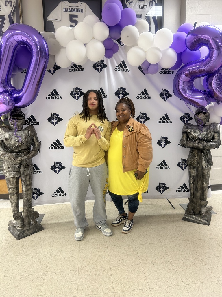 A student and an adult stand together smiling in front of a decorated Decision Day backdrop with balloons and knight statues. The student poses with a hand sign while both face the camera.